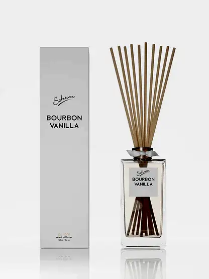 A glass bottle of "Sohum Fragrance Diffuser" by Flowers on Norton St is displayed alongside its matching rectangular box, showcasing several long, slender natural fiber reeds. The bottle contains a clear liquid infused with a refined fragrance. Both items are elegantly set against a plain white background.