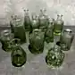 A set of distinctive glass bottles, tinted green and clear, with intricate patterns and unique shapes resembling the Vintage Vase set by Flowers on Norton St, arranged on a gray surface against a textured gray background.