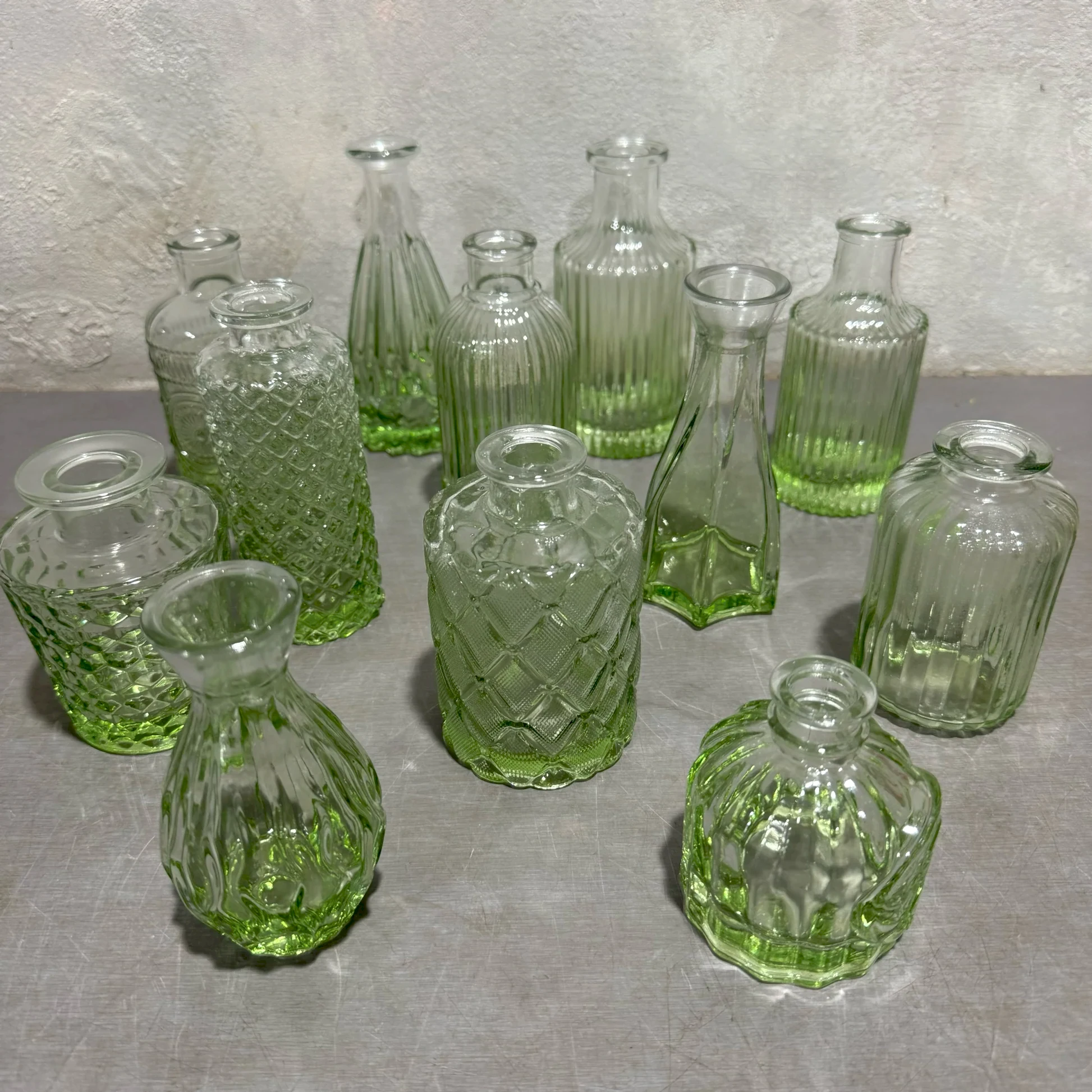 A set of distinctive glass bottles, tinted green and clear, with intricate patterns and unique shapes resembling the Vintage Vase set by Flowers on Norton St, arranged on a gray surface against a textured gray background.