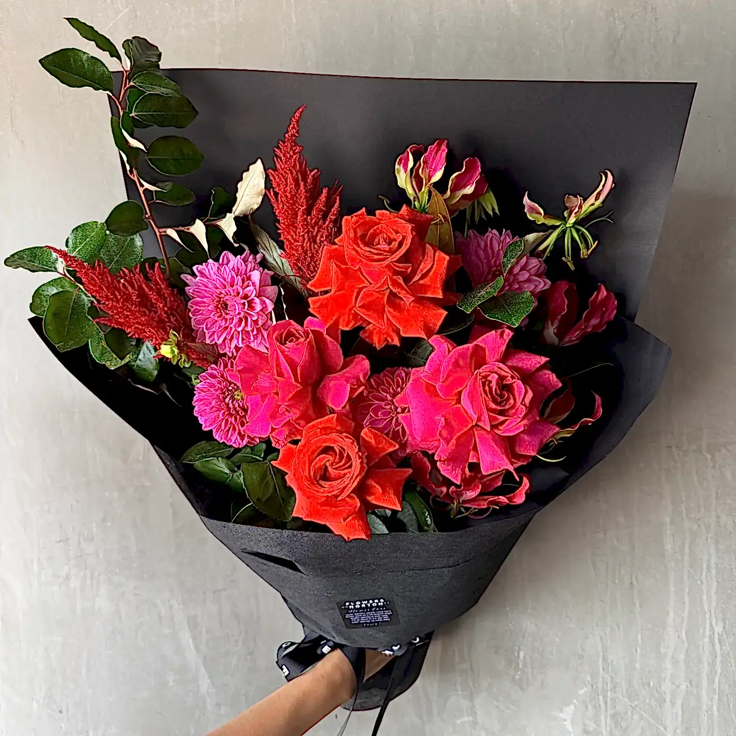 A bouquet featuring FlowersonNortonSt's Florist Choice - Red Nina Bouquet with vibrant Red Nina and pink roses, pink dahlias, and seasonal blooms with green leaves, elegantly wrapped in black paper against a gray background.