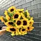 A bouquet of Sunny Sunflowers from Flowers on Norton St is held against a dark gray brick wall. The vibrant sunflowers, featuring their rich brown centers and bright green leaves, create a striking contrast with the background.