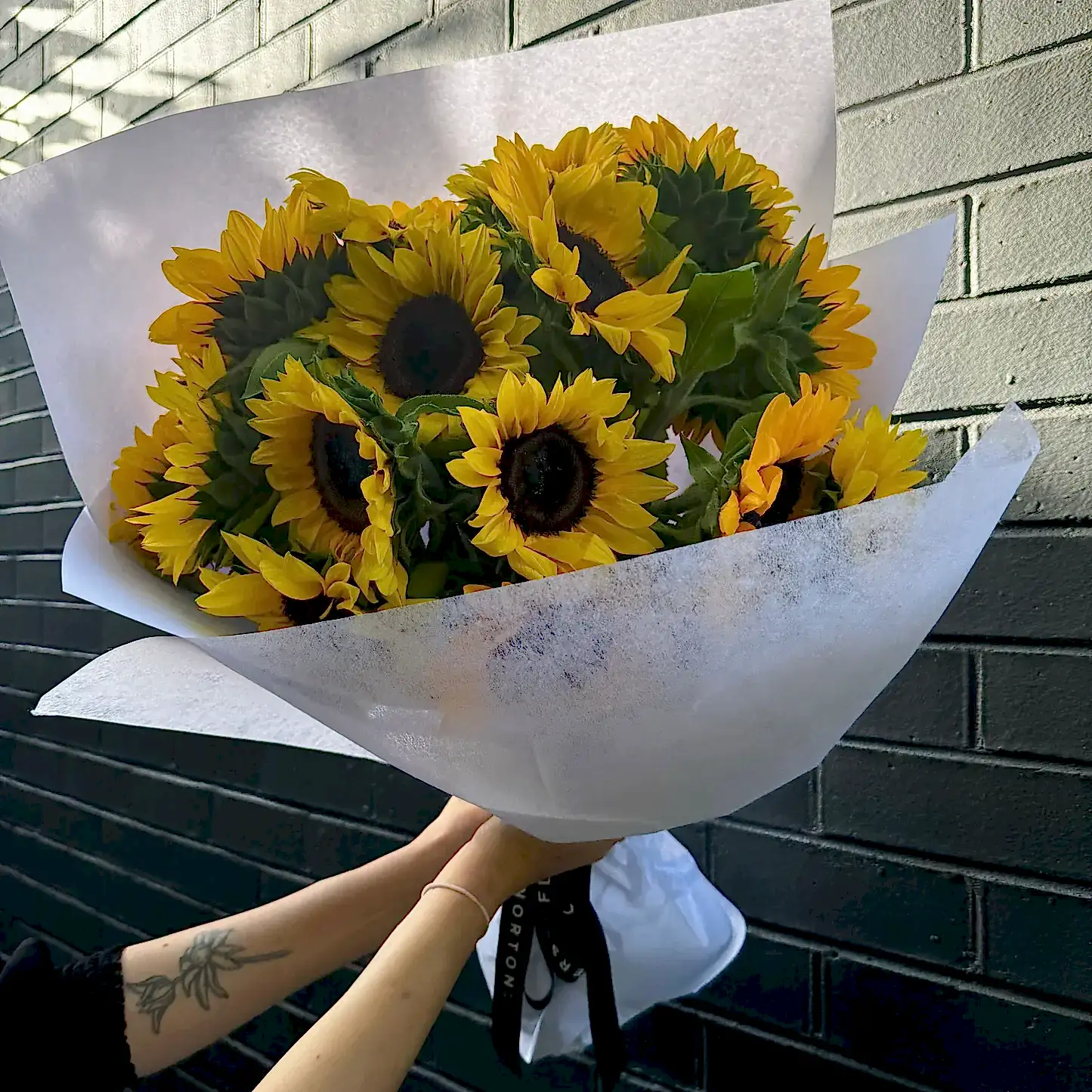A bouquet of "Sunny Sunflowers" by Flowers on Norton St is wrapped in white paper and held against a dark brick wall. The bright yellow petals provide a striking contrast to the black centers of the flowers. A tattoo adorns the arm holding this captivating arrangement.