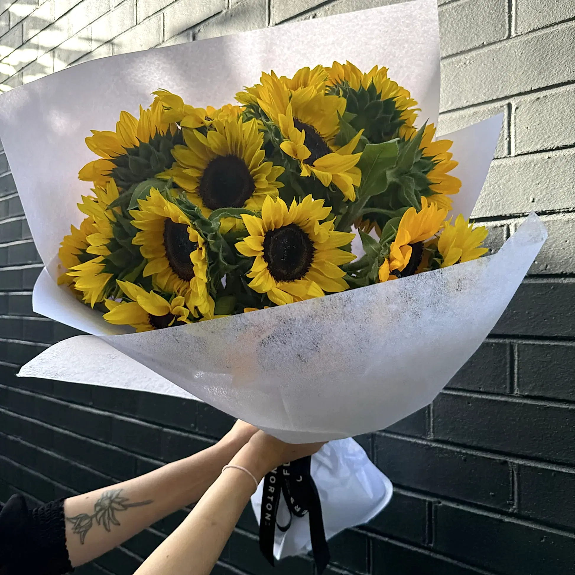 A bouquet of "Sunny Sunflowers" by Flowers on Norton St is wrapped in white paper and held against a dark brick wall. The bright yellow petals provide a striking contrast to the black centers of the flowers. A tattoo adorns the arm holding this captivating arrangement.