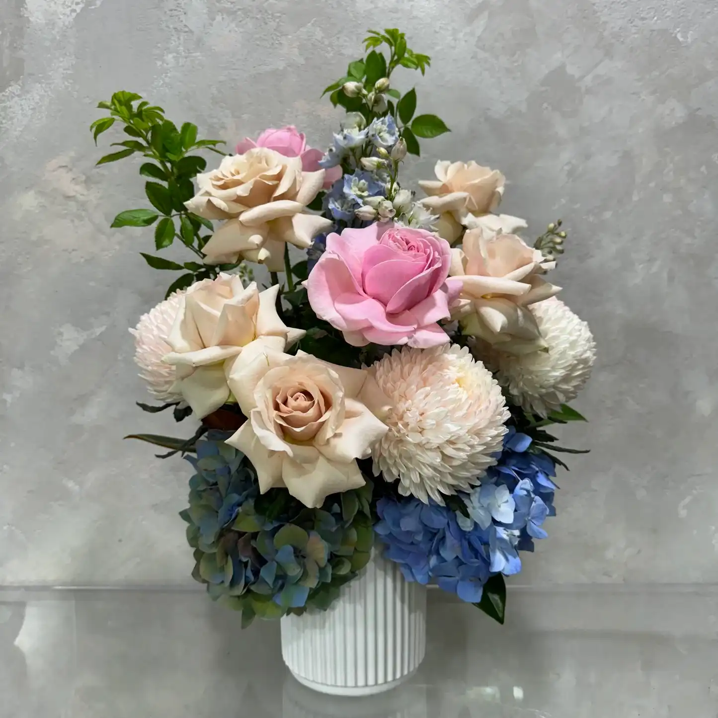 Flowers on Norton St's Mum's Garden Medley showcases a beautiful floral arrangement in a ceramic pot, highlighting pink and cream roses, white chrysanthemums, and blue hydrangeas. Set against a textured gray background, it is perfect as a Mother's Day gift.