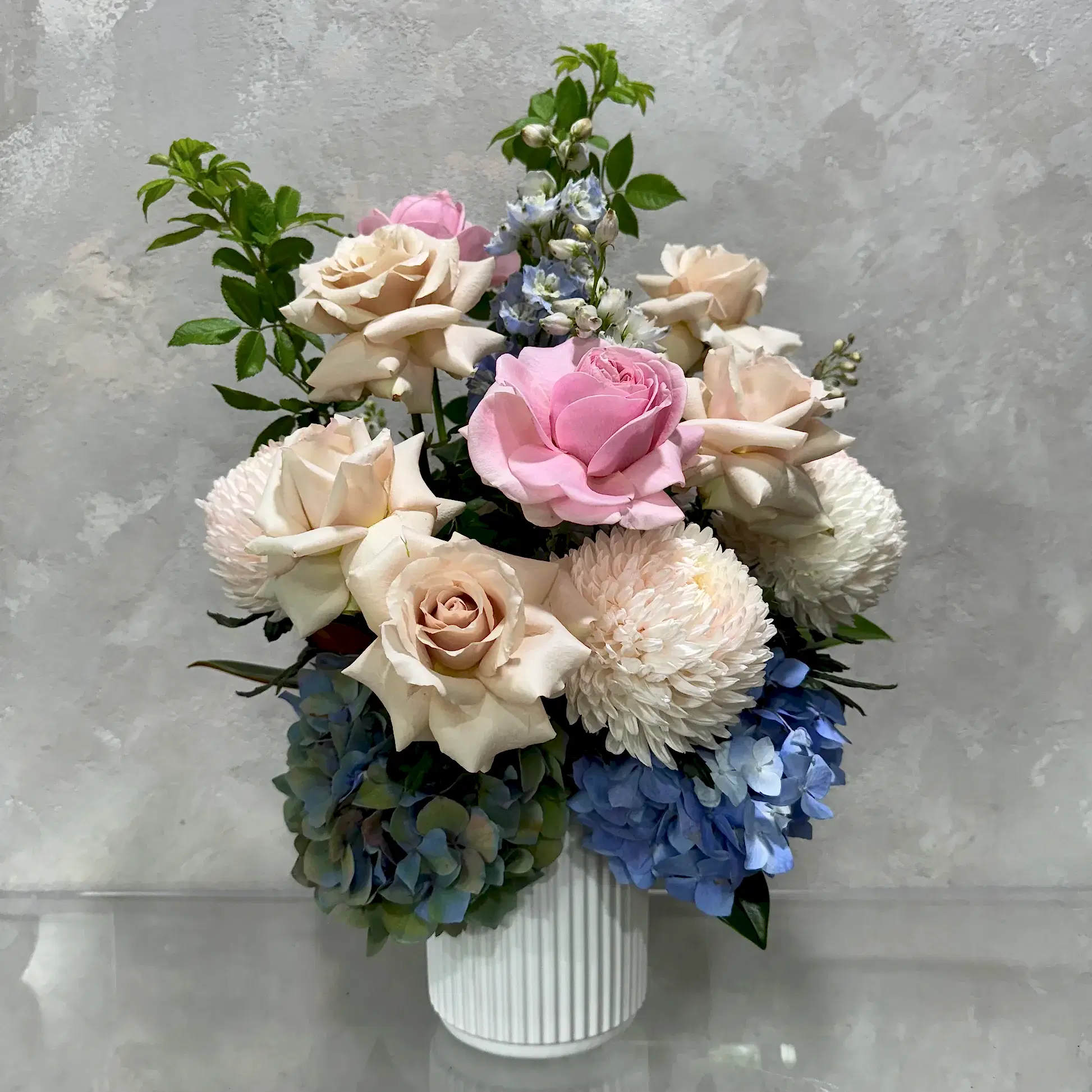 Flowers on Norton St's Mum's Garden Medley showcases a beautiful floral arrangement in a ceramic pot, highlighting pink and cream roses, white chrysanthemums, and blue hydrangeas. Set against a textured gray background, it is perfect as a Mother's Day gift.