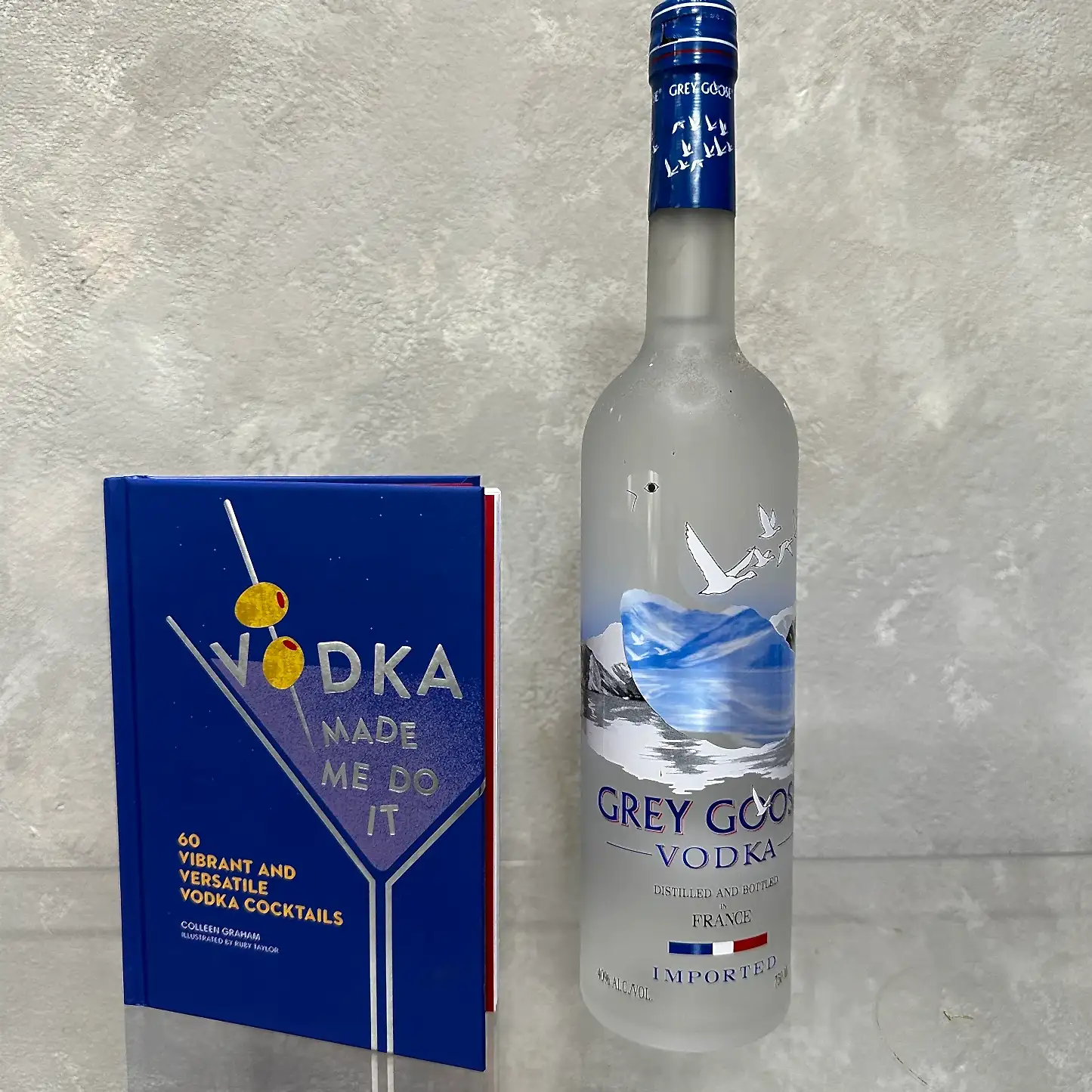 A bottle of Grey Goose vodka stands next to a blue cocktail book from the "Vodka made me do it!" Gift Set by Flowers on Norton St, adorned with images of two olives on a skewer. Both items rest against a textured, light-colored background, inviting you to explore the art of mixology.