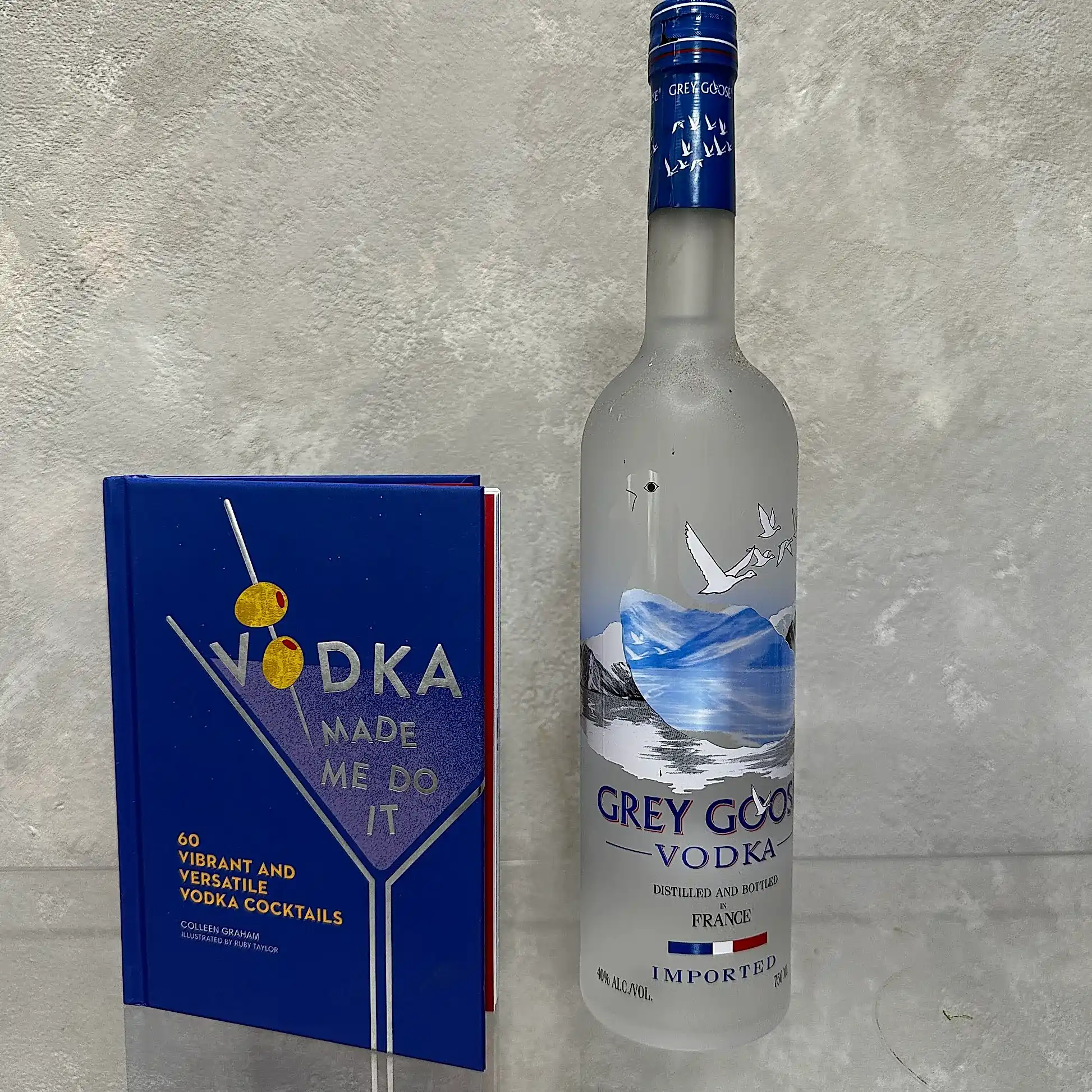 A bottle of Grey Goose vodka stands next to a blue cocktail book from the "Vodka made me do it!" Gift Set by Flowers on Norton St, adorned with images of two olives on a skewer. Both items rest against a textured, light-colored background, inviting you to explore the art of mixology.