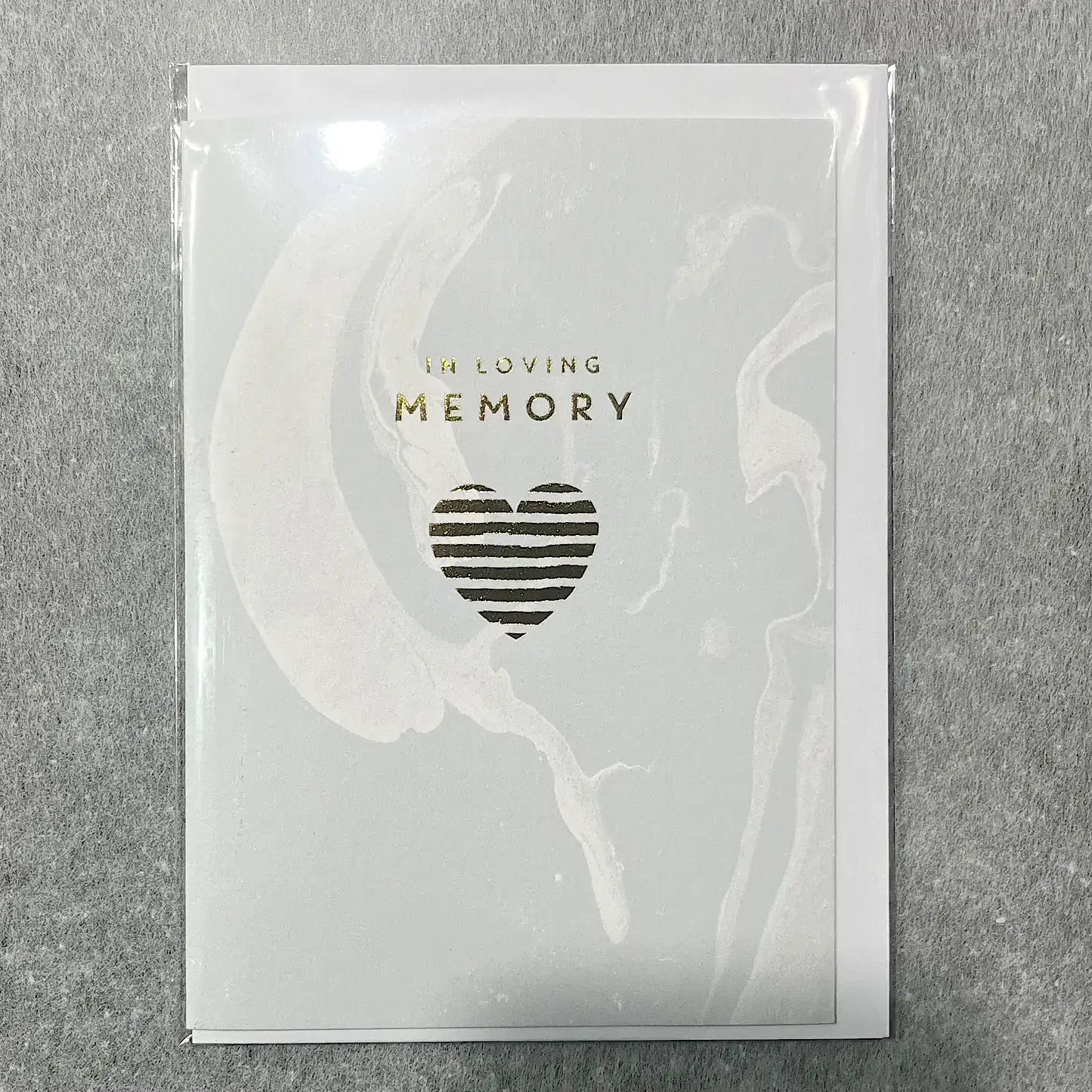 Introducing the Sympathy Cards by Flowers on Norton St: These elegant cards feature a gentle light gray background with a marbled design. The front displays the text "In Loving Memory" in shimmering gold above a heart-shaped stripe. Each card is thoughtfully crafted and enclosed in a clear plastic cover, making it an ideal addition to any florist's collection.