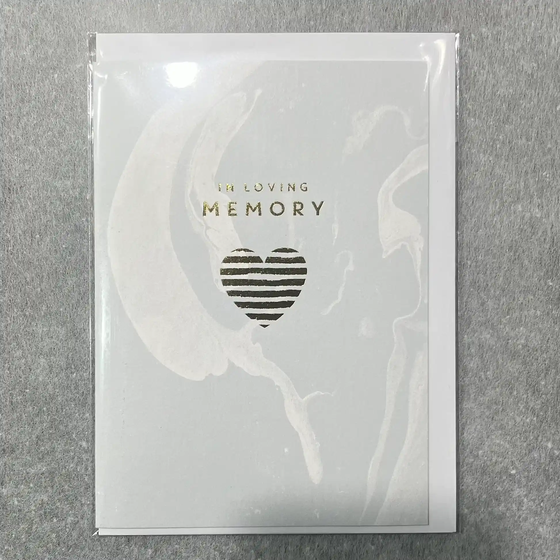 Introducing the Sympathy Cards by Flowers on Norton St: These elegant cards feature a gentle light gray background with a marbled design. The front displays the text "In Loving Memory" in shimmering gold above a heart-shaped stripe. Each card is thoughtfully crafted and enclosed in a clear plastic cover, making it an ideal addition to any florist's collection.