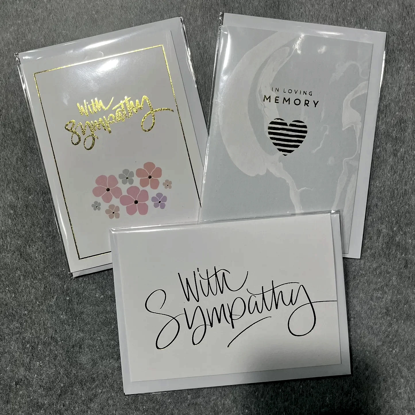 Displayed on a gray surface are three sympathy cards from Flowers on Norton St. One card features pink flowers with gold lettering, another showcases a heart design with the words "IN LOVING MEMORY," and the third is adorned with elegant black script reading "With Sympathy.