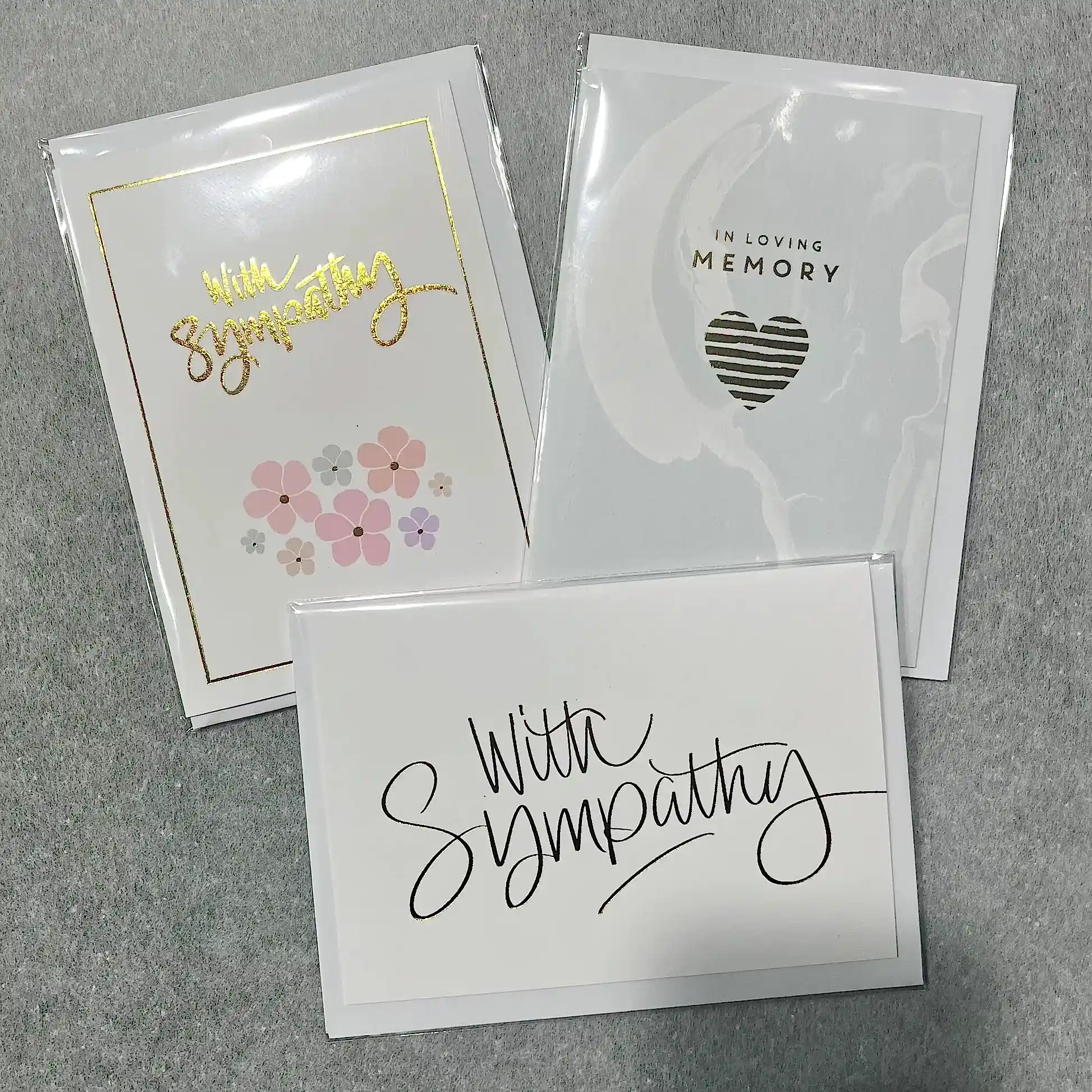 Displayed on a gray surface are three sympathy cards from Flowers on Norton St. One card features pink flowers with gold lettering, another showcases a heart design with the words "IN LOVING MEMORY," and the third is adorned with elegant black script reading "With Sympathy.
