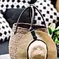 A straw hat, fastened with an accessory from Klipsta and featuring a black band, is placed on a woven basket with leather handles. The basket rests on a table, complemented by stylish black and white patterned cushions in the background. Lush plants from the side enhance the scene with a touch of nature. This beautiful arrangement showcases products by Flowers on Norton St.