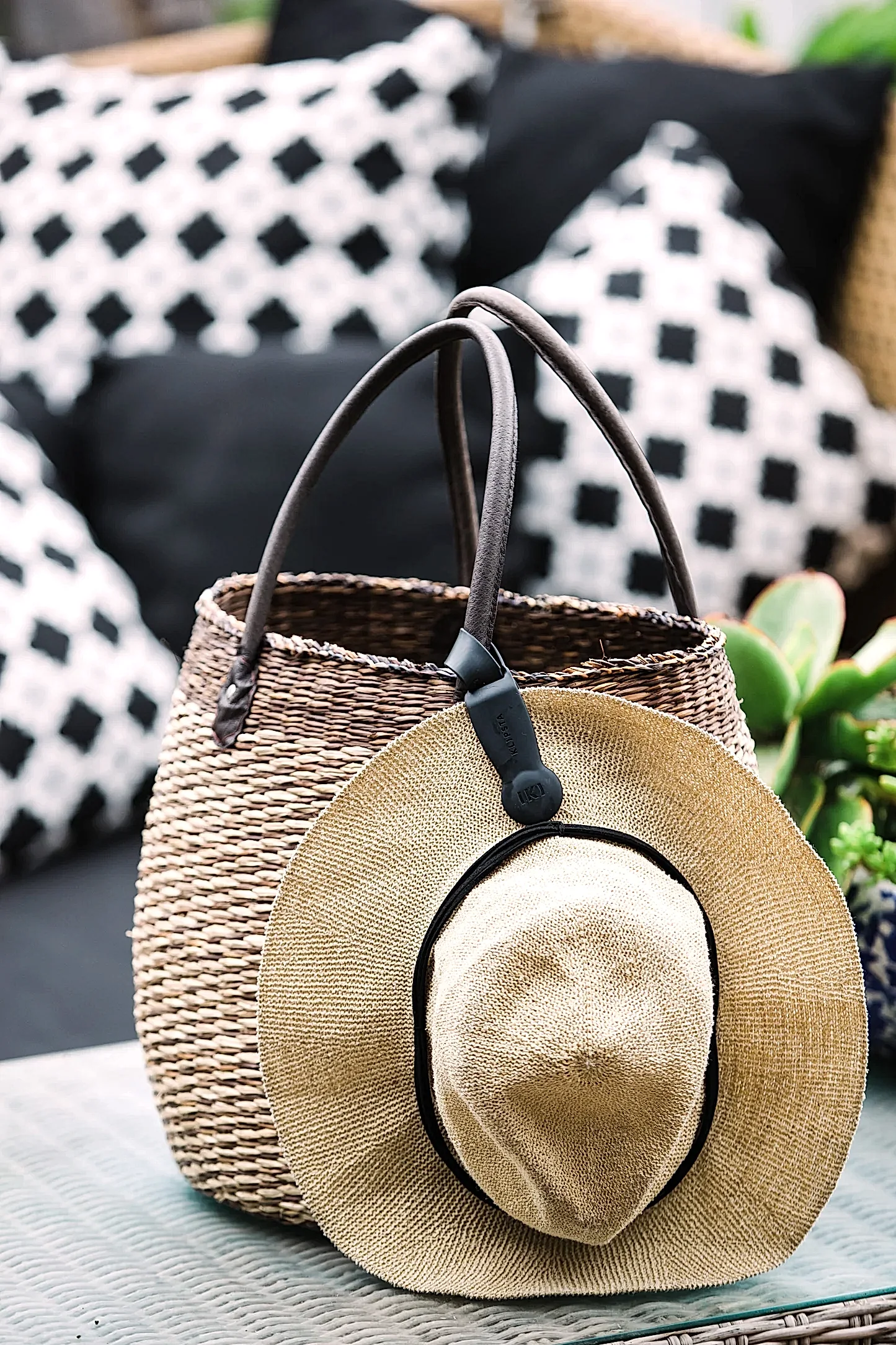 A straw hat, fastened with an accessory from Klipsta and featuring a black band, is placed on a woven basket with leather handles. The basket rests on a table, complemented by stylish black and white patterned cushions in the background. Lush plants from the side enhance the scene with a touch of nature. This beautiful arrangement showcases products by Flowers on Norton St.
