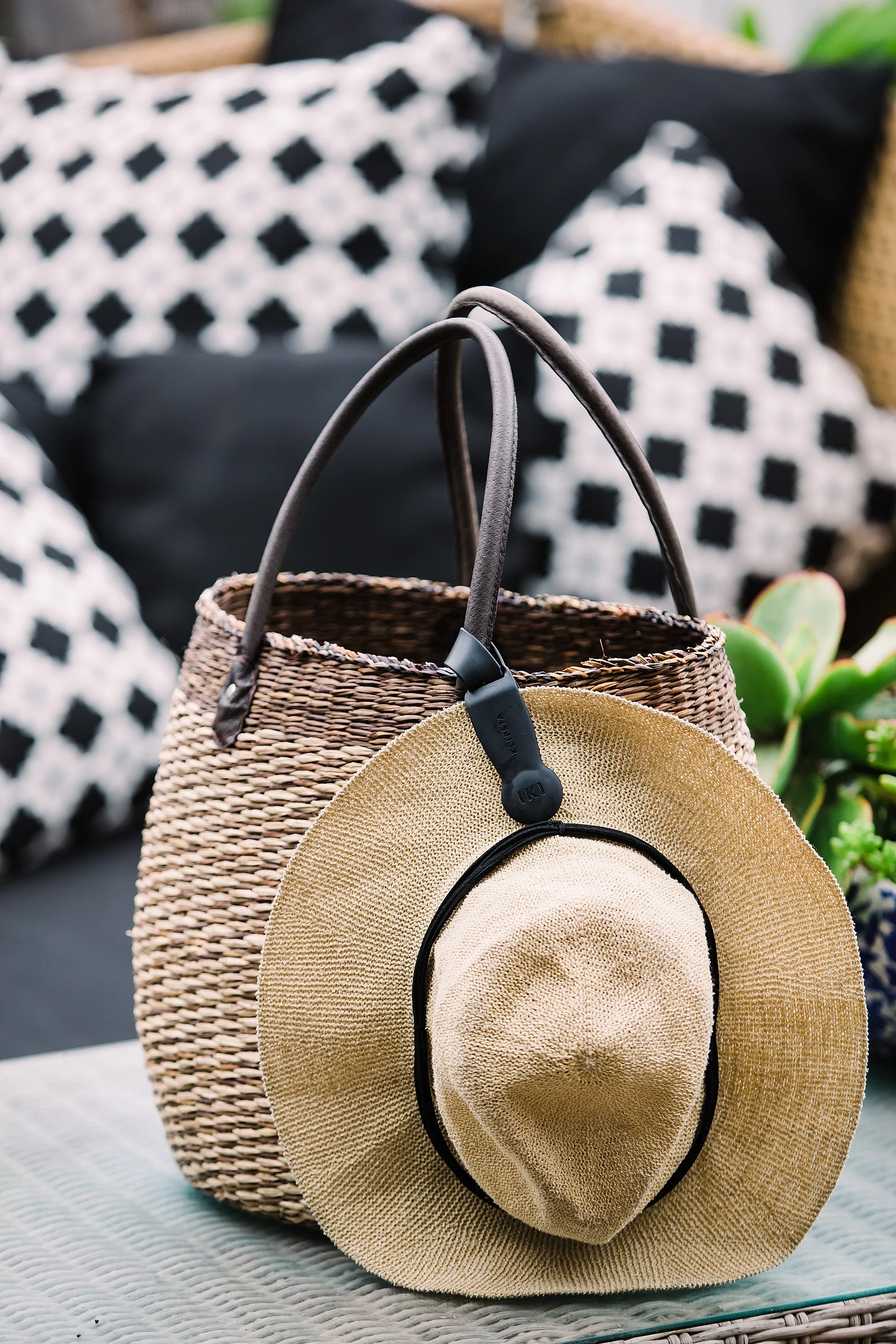 A straw hat, fastened with an accessory from Klipsta and featuring a black band, is placed on a woven basket with leather handles. The basket rests on a table, complemented by stylish black and white patterned cushions in the background. Lush plants from the side enhance the scene with a touch of nature. This beautiful arrangement showcases products by Flowers on Norton St.
