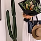 A person in a white dress holds a bouquet from Flowers on Norton St, wrapped in black paper. Using an Accessories by Klipsta hat clip, their tan hat hangs neatly from a black tote bag. A tall, green cactus stands beside them against a white wall.