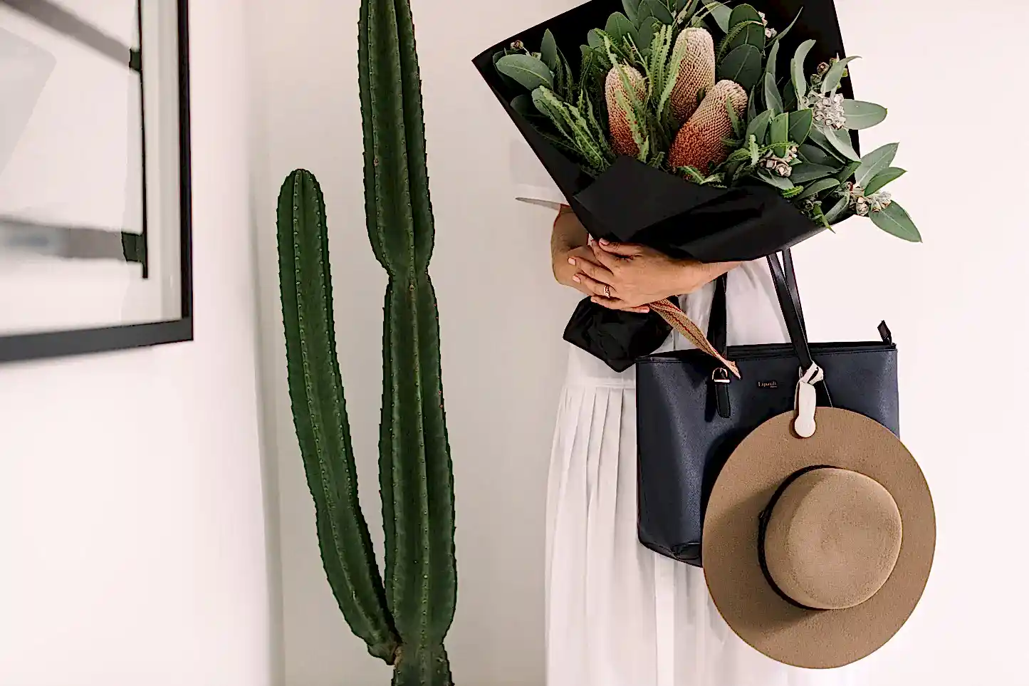 A person in a white dress holds a bouquet from Flowers on Norton St, wrapped in black paper. Using an Accessories by Klipsta hat clip, their tan hat hangs neatly from a black tote bag. A tall, green cactus stands beside them against a white wall.