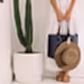 A person in a white dress is depicted holding a navy bag and a wide-brimmed brown hat fastened with an Accessories by Klipsta clip from Flowers on Norton St. They are standing beside a tall cactus in a white planter, wearing sandals. The scene presents minimalist aesthetics featuring neutral tones.