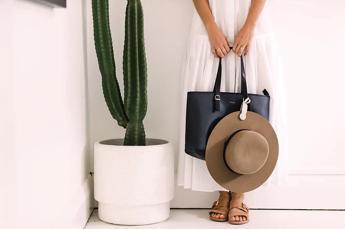 A person in a white dress is depicted holding a navy bag and a wide-brimmed brown hat fastened with an Accessories by Klipsta clip from Flowers on Norton St. They are standing beside a tall cactus in a white planter, wearing sandals. The scene presents minimalist aesthetics featuring neutral tones.