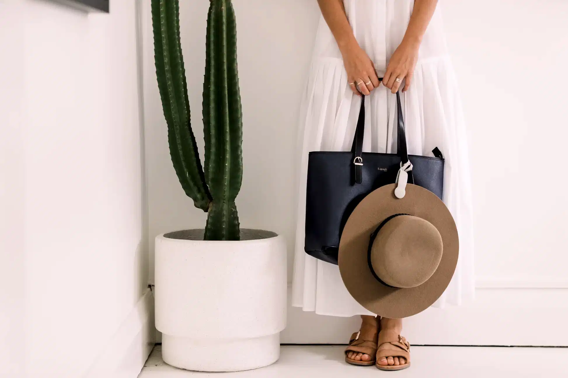 A person in a white dress is depicted holding a navy bag and a wide-brimmed brown hat fastened with an Accessories by Klipsta clip from Flowers on Norton St. They are standing beside a tall cactus in a white planter, wearing sandals. The scene presents minimalist aesthetics featuring neutral tones.