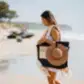 A woman strolls down the sandy beach, her black tote adorned with Accessories by Klipsta from Flowers on Norton St securing her straw hat. Dressed in a white outfit and a light scarf draped over her shoulder, she is surrounded by blurred trees and a nearby jet ski near the shoreline.