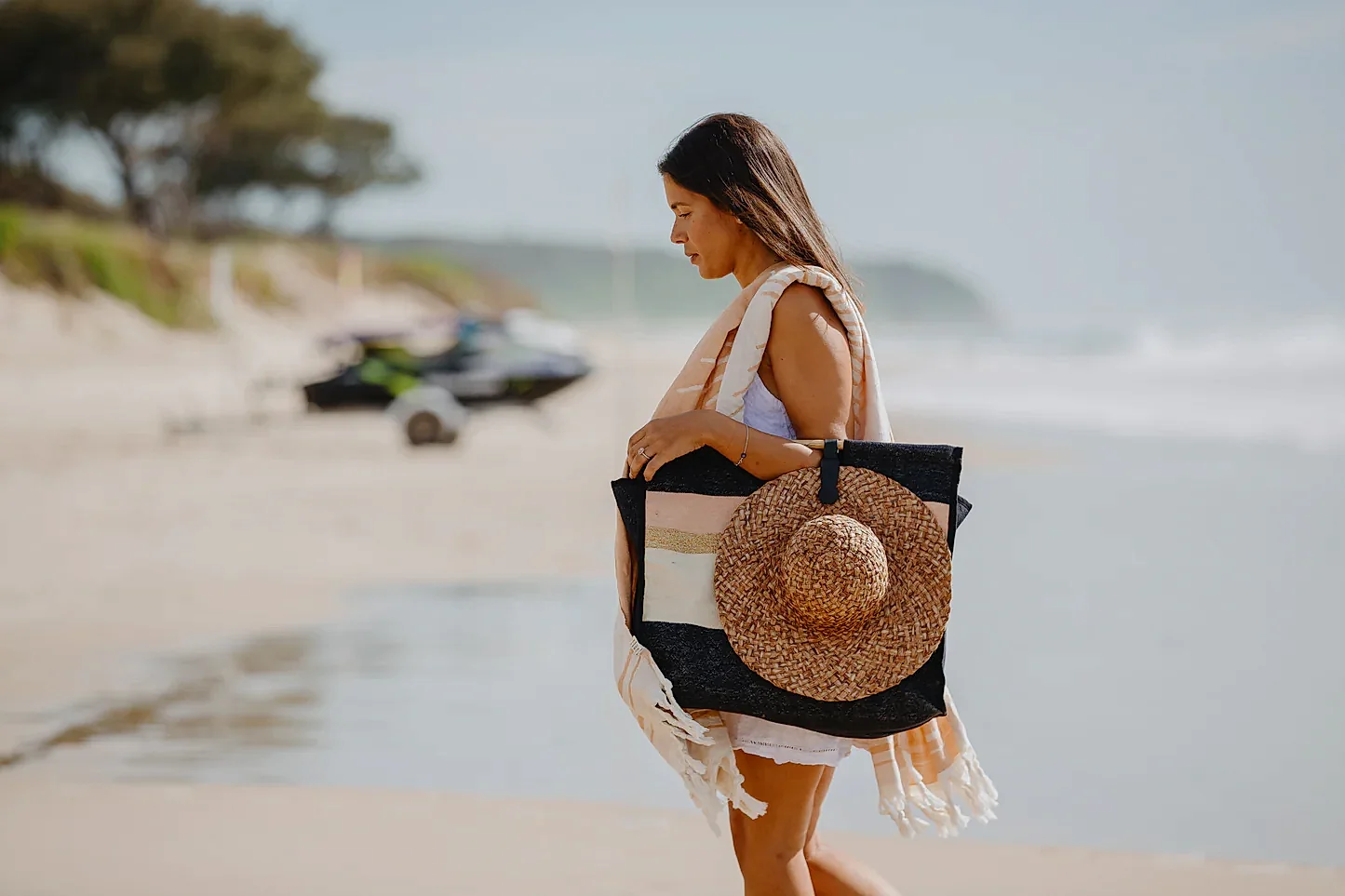 A woman strolls down the sandy beach, her black tote adorned with Accessories by Klipsta from Flowers on Norton St securing her straw hat. Dressed in a white outfit and a light scarf draped over her shoulder, she is surrounded by blurred trees and a nearby jet ski near the shoreline.