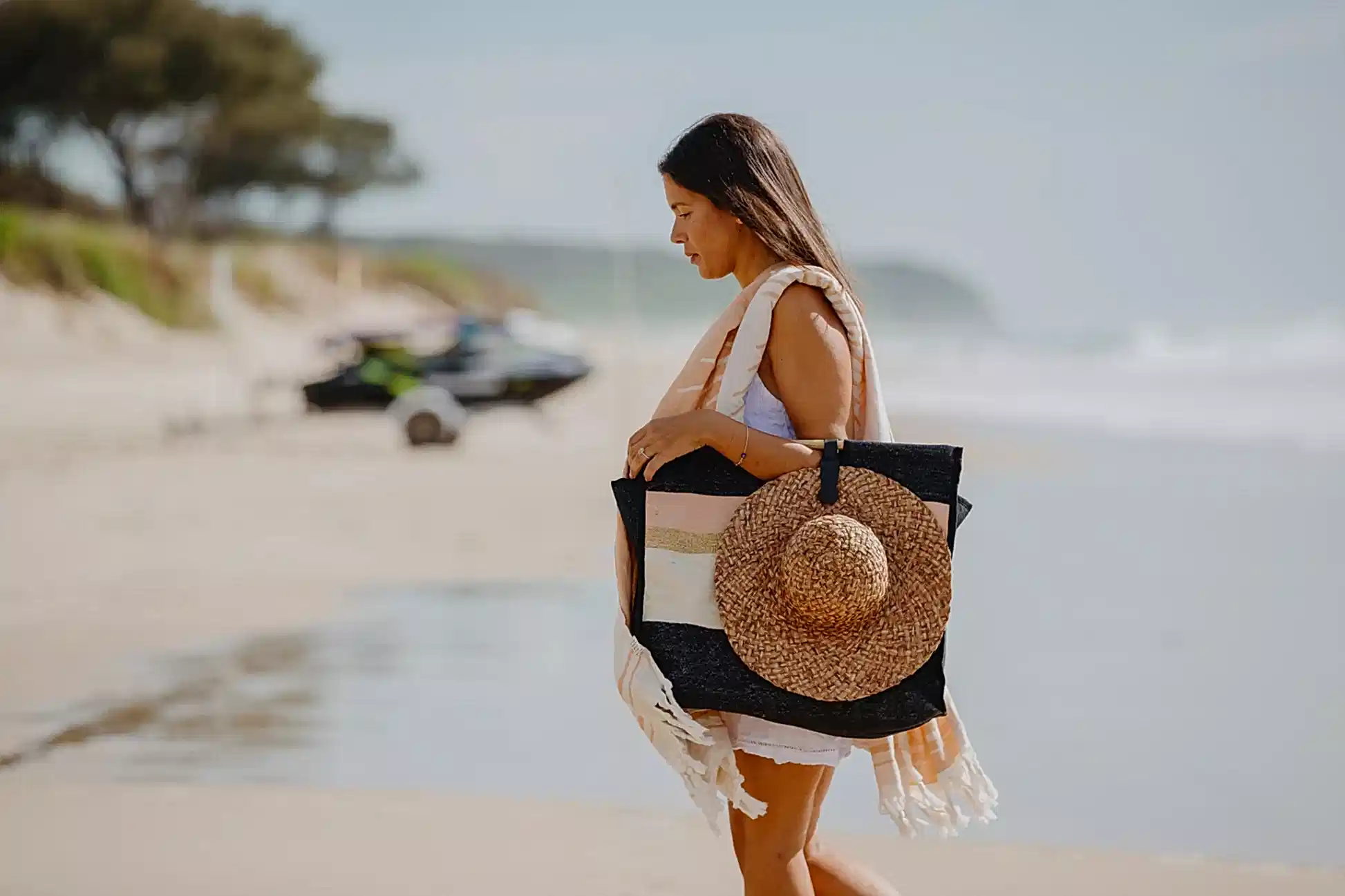 A woman strolls down the sandy beach, her black tote adorned with Accessories by Klipsta from Flowers on Norton St securing her straw hat. Dressed in a white outfit and a light scarf draped over her shoulder, she is surrounded by blurred trees and a nearby jet ski near the shoreline.
