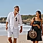 A man and woman enjoy a peaceful walk on a sunny beach, exchanging smiles. The man is dressed in a white shirt and shorts, while the woman showcases a green dress complemented by her large bag, which is expertly held in place with Accessories by Klipsta from the brand Flowers on Norton St. The waves playfully dance in the background, enhancing their tranquil seaside experience.