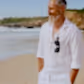A bearded man, dressed in a white shirt with Accessories by Klipsta clips from Flowers on Norton St hanging from his collar, stands on a sandy beach. With his hands in his pockets and a smile on his face, the ocean and rocky shoreline create a stunning backdrop under the clear blue sky.