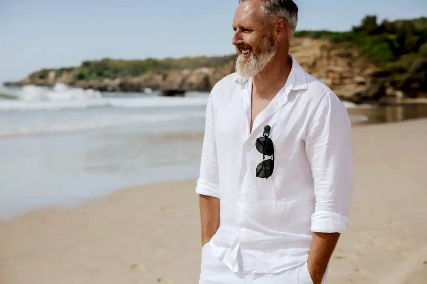 A bearded man, dressed in a white shirt with Accessories by Klipsta clips from Flowers on Norton St hanging from his collar, stands on a sandy beach. With his hands in his pockets and a smile on his face, the ocean and rocky shoreline create a stunning backdrop under the clear blue sky.