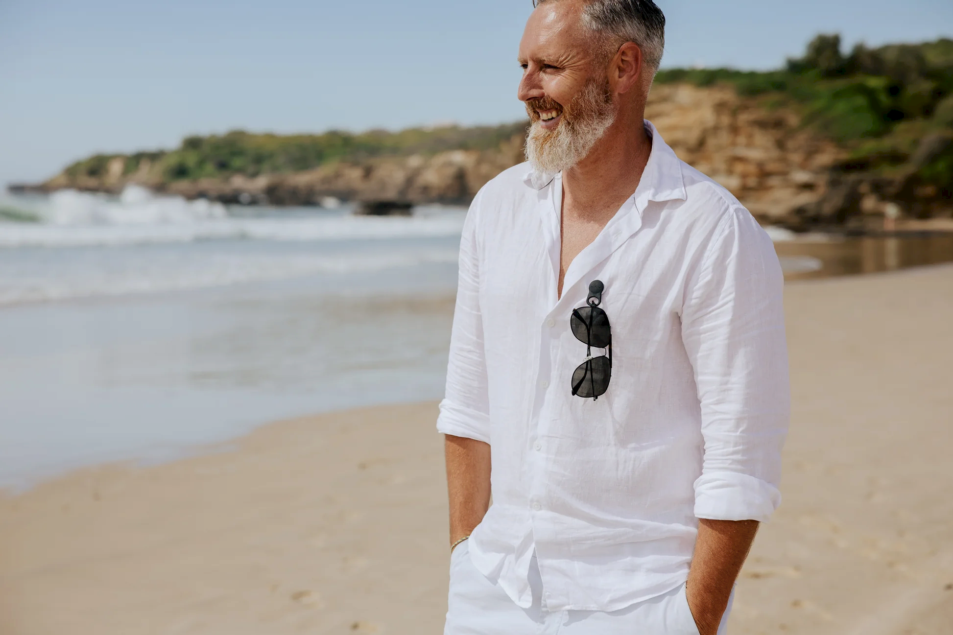 A bearded man, dressed in a white shirt with Accessories by Klipsta clips from Flowers on Norton St hanging from his collar, stands on a sandy beach. With his hands in his pockets and a smile on his face, the ocean and rocky shoreline create a stunning backdrop under the clear blue sky.