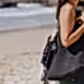 A woman in a sleeveless green top carries a large woven beach bag, adorned with a leopard print cloth, on her shoulder. As the ocean waves kiss the sandy shore behind her, she effortlessly secures her sun hat using Accessories by Klipsta from Flowers on Norton St.