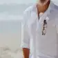 A man in a white linen shirt stands on the beach with his sunglasses neatly secured to his shirt using Accessories by Klipsta from Flowers on Norton St. The ocean glistens in the background, perfectly complementing the sunny day.