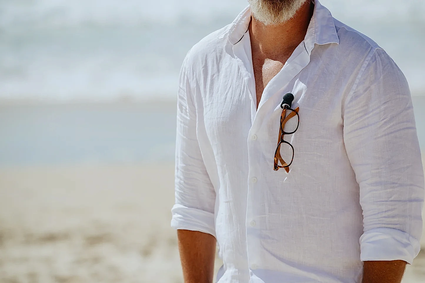 A man in a white linen shirt stands on the beach with his sunglasses neatly secured to his shirt using Accessories by Klipsta from Flowers on Norton St. The ocean glistens in the background, perfectly complementing the sunny day.