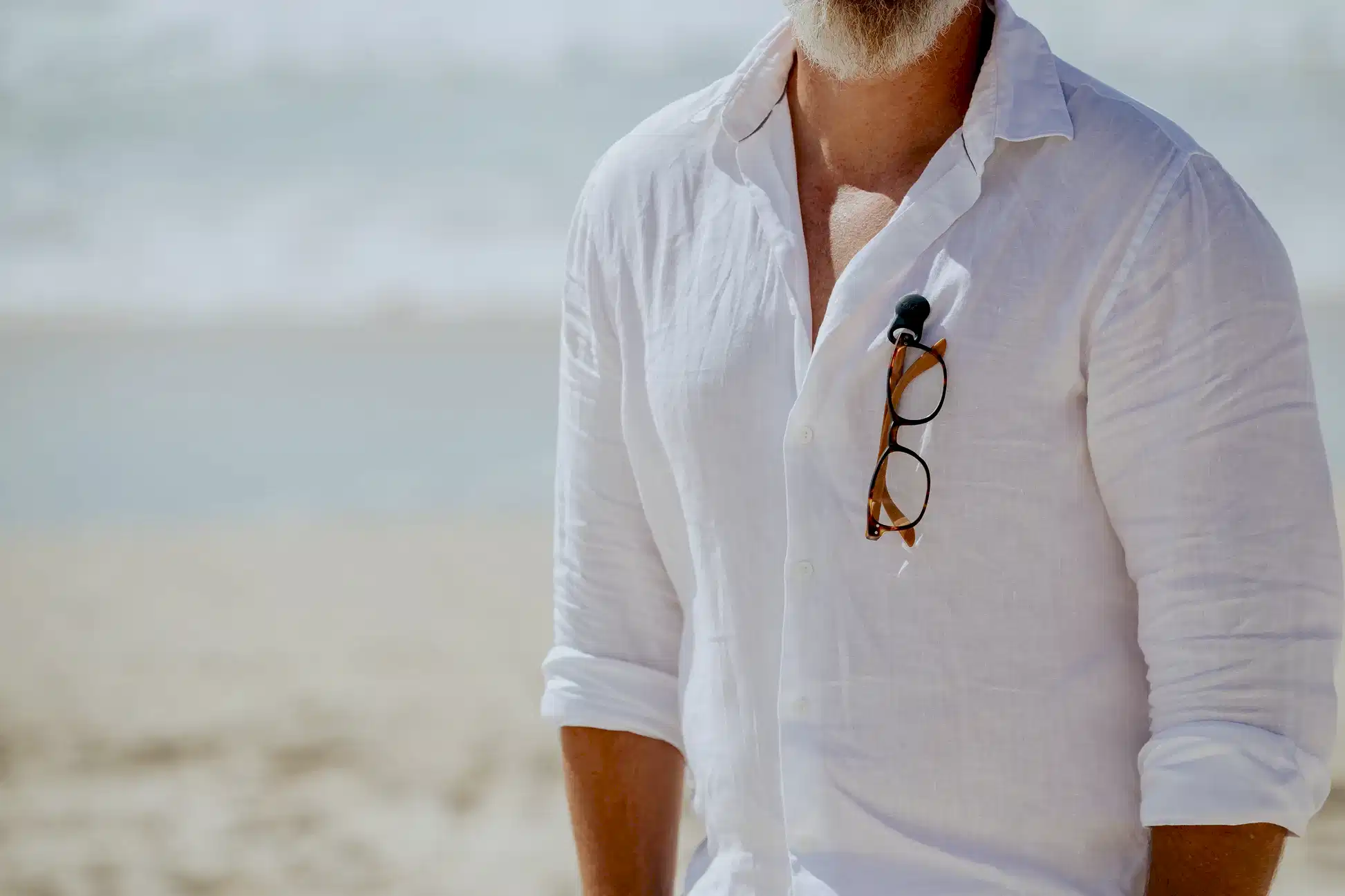 A man in a white linen shirt stands on the beach with his sunglasses neatly secured to his shirt using Accessories by Klipsta from Flowers on Norton St. The ocean glistens in the background, perfectly complementing the sunny day.