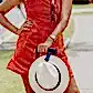 A woman wearing a sleeveless orange dress is outdoors on a sunny day in a suburban area, holding the handle of a black suitcase. She has sunglasses neatly secured at her neckline with Accessories by Klipsta from Flowers on Norton St and her white hat is smartly attached to the suitcase using hat clips from the same brand.
