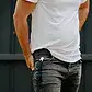 A person wearing a white t-shirt and black jeans stands with their hand in their pocket, where a pair of sunglasses and an Accessories by Klipsta AirPods case from Flowers on Norton St are securely clipped. The backdrop is a dark wall.
