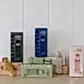 Five Accessories by Klipsta magnetic hat clips from Flowers on Norton St are showcased in vibrant colors—green, blue, beige, and pink—against a white wall. The clips are creatively arranged on and around two cardboard boxes, forming a simple yet minimalist display.