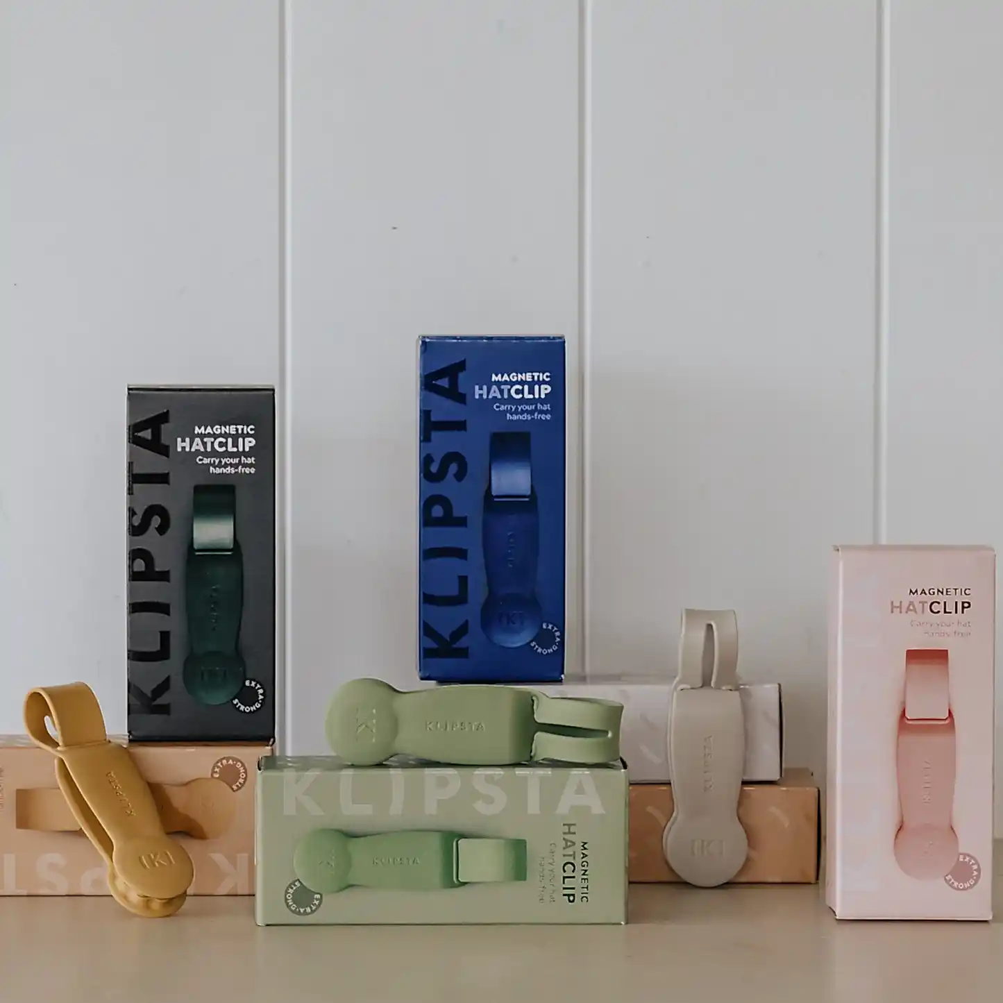 Five Accessories by Klipsta magnetic hat clips from Flowers on Norton St are showcased in vibrant colors—green, blue, beige, and pink—against a white wall. The clips are creatively arranged on and around two cardboard boxes, forming a simple yet minimalist display.