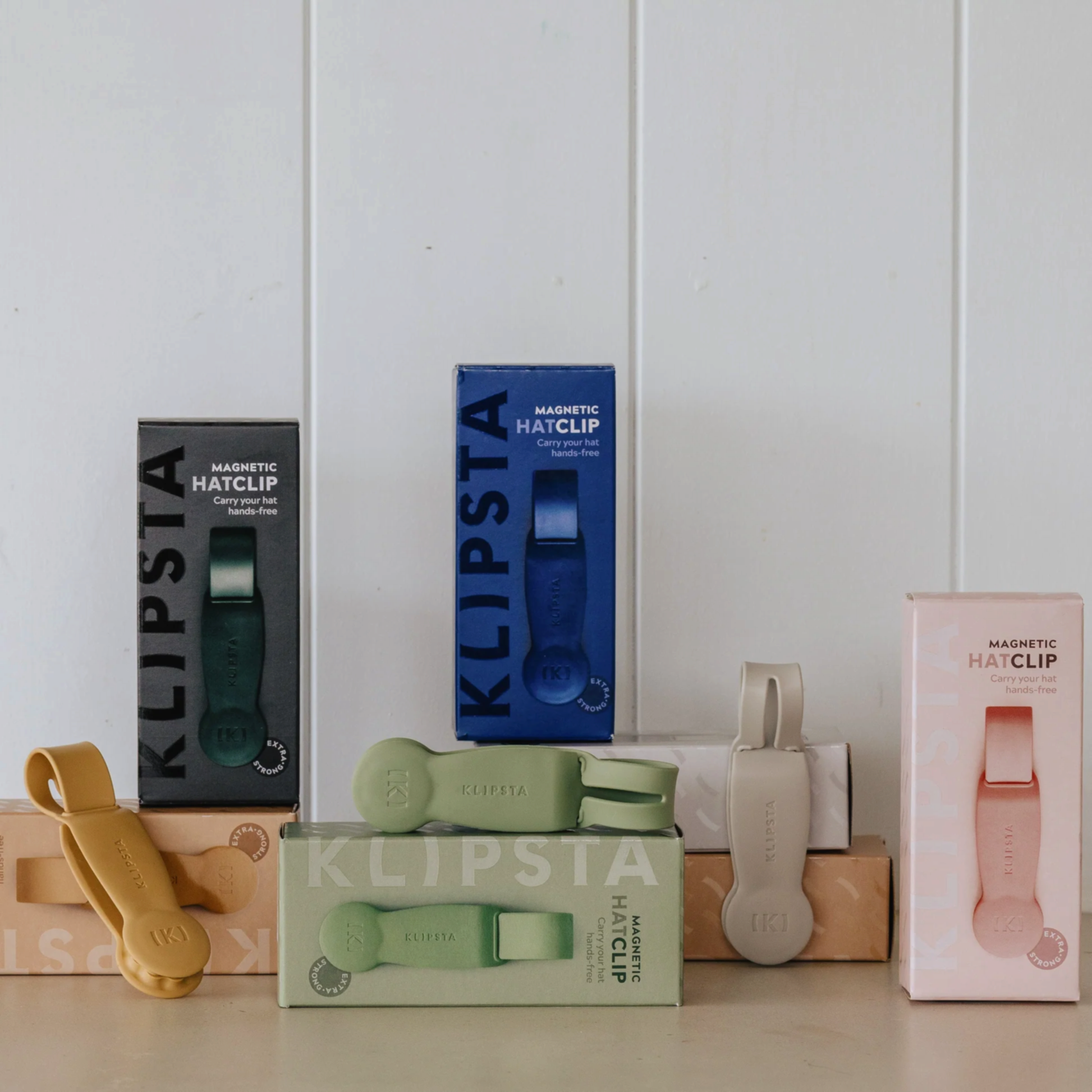 Five Accessories by Klipsta magnetic hat clips from Flowers on Norton St are showcased in vibrant colors—green, blue, beige, and pink—against a white wall. The clips are creatively arranged on and around two cardboard boxes, forming a simple yet minimalist display.