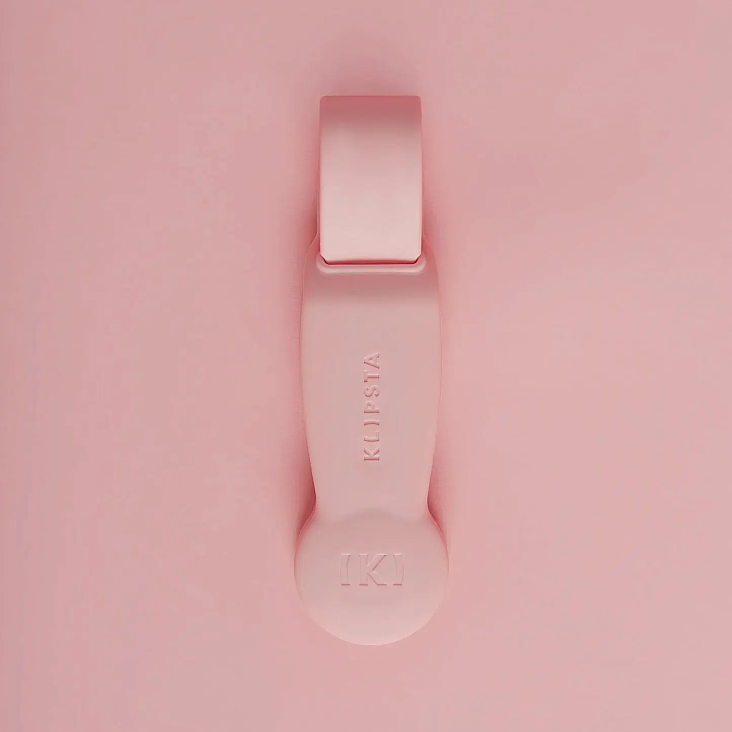 Against a matching pink background lies a flat tool with a looped handle and rounded end, embossed with the logo "KLIPSTA." This versatile accessory, part of the "Accessories by Klipsta" collection from Flowers on Norton St, is perfect for stylishly clipping glasses or hats.