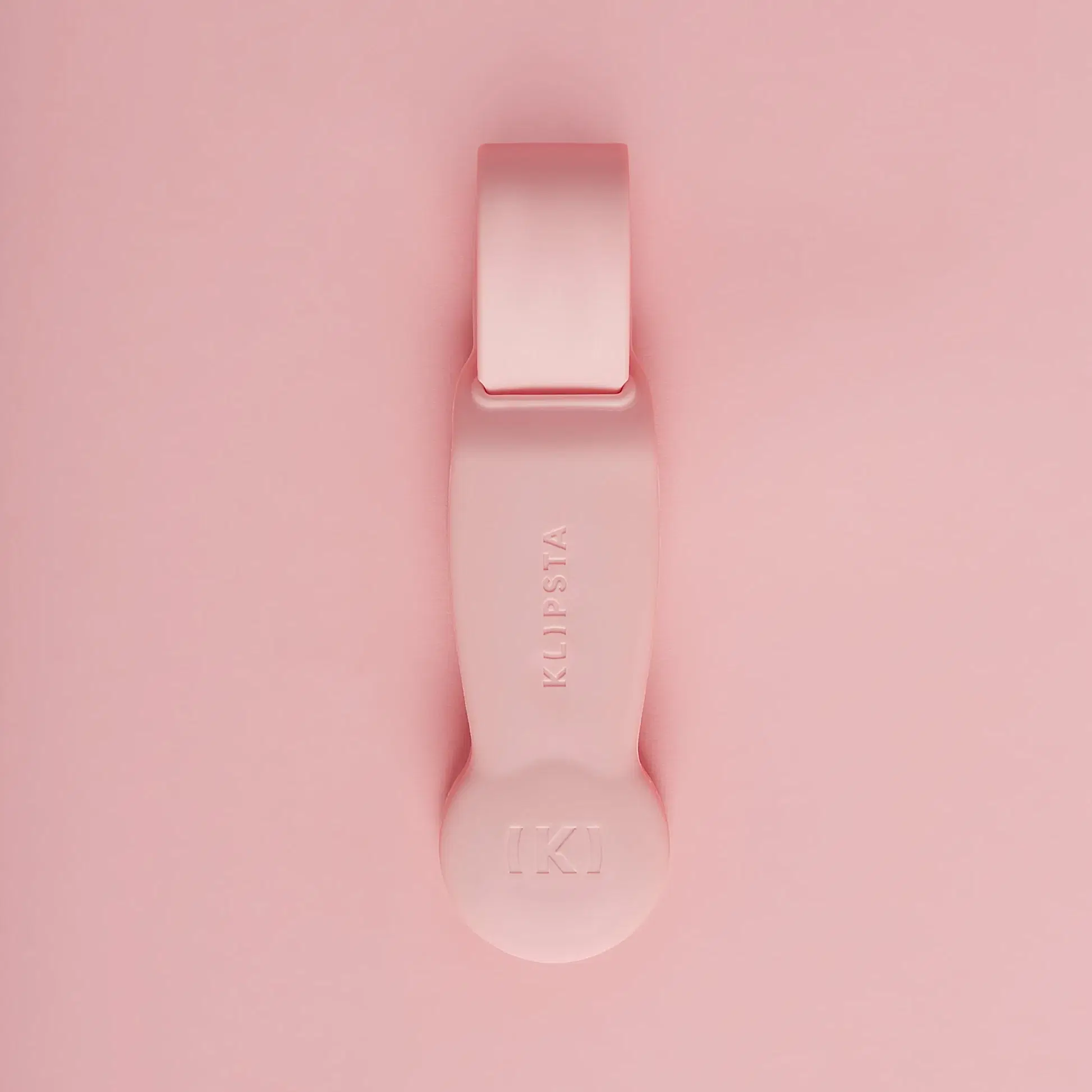 Against a matching pink background lies a flat tool with a looped handle and rounded end, embossed with the logo "KLIPSTA." This versatile accessory, part of the "Accessories by Klipsta" collection from Flowers on Norton St, is perfect for stylishly clipping glasses or hats.