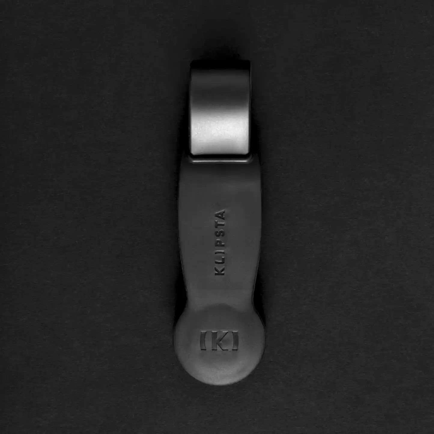 A sophisticated black hat clip from the "Accessories by Klipsta" collection, featuring an elegant design, prominently showcasing the name "KLIPSTA" and a raised "KI" logo, all gracefully contrasted against a dark backdrop, crafted by Flowers on Norton St.