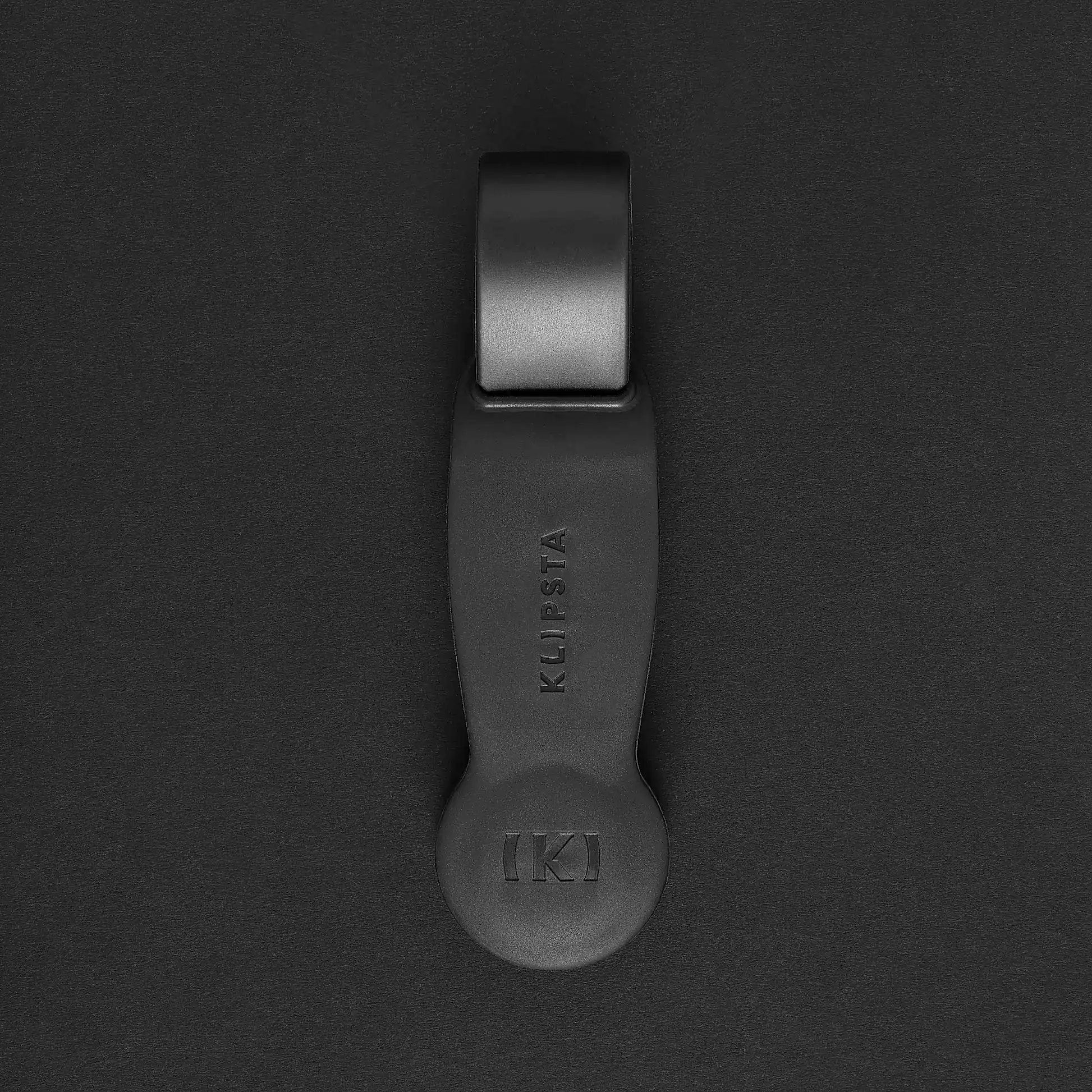 A sophisticated black hat clip from the "Accessories by Klipsta" collection, featuring an elegant design, prominently showcasing the name "KLIPSTA" and a raised "KI" logo, all gracefully contrasted against a dark backdrop, crafted by Flowers on Norton St.