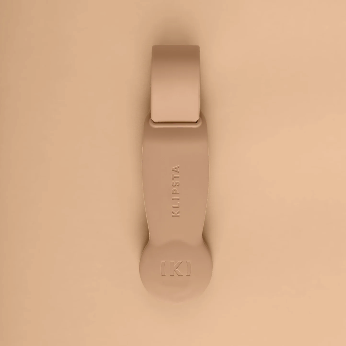 A beige, minimalist accessory from Flowers on Norton St set against a matching background. This oblong piece with a rounded bottom features "KLIPSTA" and a stylized K logo. It functions as either hat clips or glasses clips, offering a sleek, modern design that stylishly organizes your essentials.