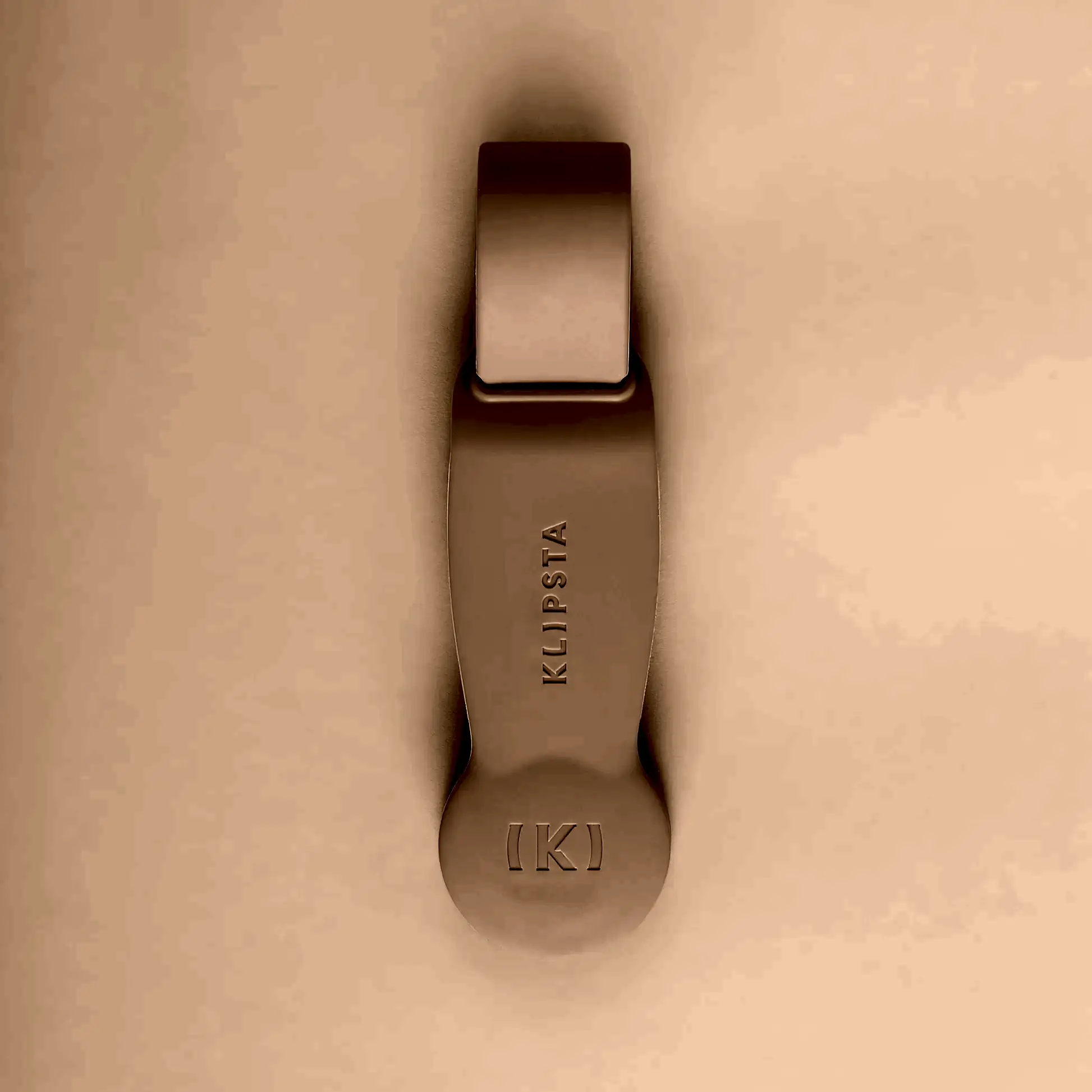 A beige, minimalist accessory from Flowers on Norton St set against a matching background. This oblong piece with a rounded bottom features "KLIPSTA" and a stylized K logo. It functions as either hat clips or glasses clips, offering a sleek, modern design that stylishly organizes your essentials.