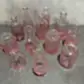 A set of charming, miniature clear glass vases with a pink tint from Flowers on Norton St's Vintage Vase collection is showcased on a gray surface against a textured wall. These elegant bottles come in diverse shapes and intricate patterns, perfect for adding a decorative flair.