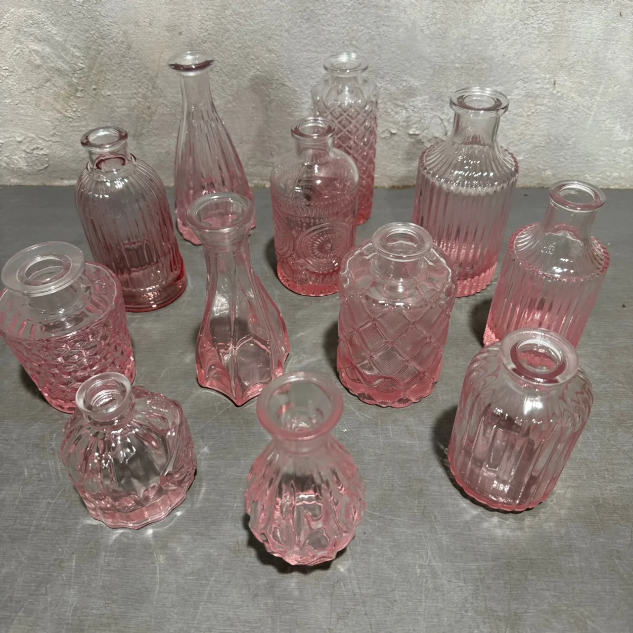 A set of charming, miniature clear glass vases with a pink tint from Flowers on Norton St's Vintage Vase collection is showcased on a gray surface against a textured wall. These elegant bottles come in diverse shapes and intricate patterns, perfect for adding a decorative flair.