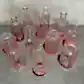 A Vintage Vase set from Flowers on Norton St is artfully arranged on a gray surface, showcasing a collection of small, intricately designed pink glass bottles and mini vases. The bottles come in various shapes and textures, featuring patterns such as ribbing and diamond cuts. A textured, light-colored wall serves as the backdrop.