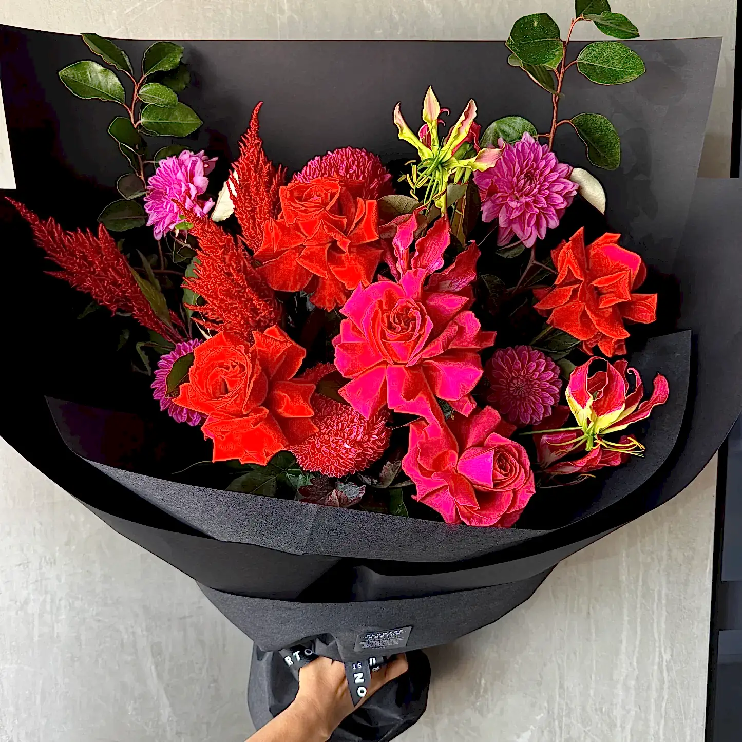 The Florist Choice - Red Nina Bouquet by FlowersonNortonSt boasts vibrant Red Nina and Colombian roses with purple dahlias and green leaves, beautifully wrapped in black paper.