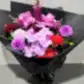 The Be Bold Seasonal Bouquet by FlowersonNortonSt showcases a vibrant mix of seasonal flowers, including pink phalaenopsis orchids, roses, dahlias, and snapdragons. This elegant arrangement is wrapped in black paper and presented on a gray surface.